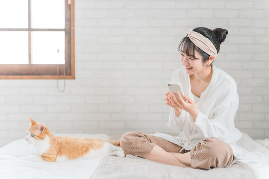 家のベッドルームでペットの猫とスマホで写真を撮って遊ぶアジア人女性

