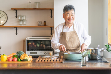 家のキッチンでフライパンを使って手料理を作るアジア人の高齢者男性（笑顔）
