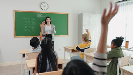 Caucasian female teacher standing at green chalkboard teaching and writing with chalk in mathematics subject at elementary school. Students raising hand answer question teacher in classroom. - Powered by Adobe