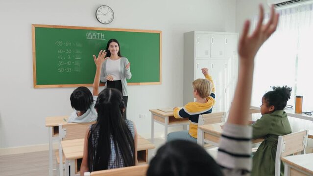 Caucasian female teacher standing at green chalkboard teaching and writing with chalk in mathematics subject at elementary school. Students raising hand answer question teacher in classroom.