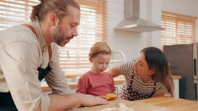 Happy family consists of father mother and little son kneading dough together in the kitchen. Caucasian parents teaching son to prepare dough for cookies and enjoying spending free time in kitchen.
