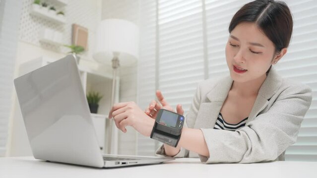 Asian business woman use blood pressure monitor on wrist checking blood pressure and heartbeat while working in office room. Young asian female health care in workplace. Healthcare and medical concept