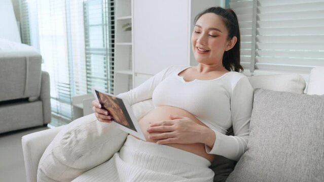 Pregnancy asian mother looking picture of ultrasound film and touching her abdomen. Asian pregnant woman checking result ultrasound baby sitting relaxed and rest on sofa in living room at home.