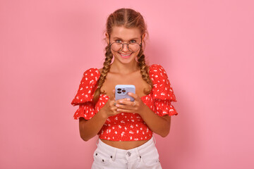 Young cheerful beautiful Caucasian woman student in glasses holding mobile phone getting distance education and studying in internet institute or college through website stands in pink studio.