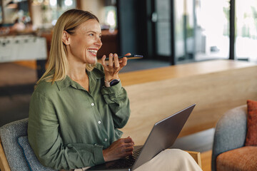 Smiling businesswoman is record audio massege on phone in coworking and work on laptop
