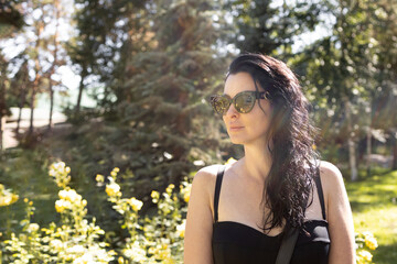 Smiling long-haired woman chilling in resort. Beautiful brunette dressed in black fitting dress and sunglasses walking in the park. Chasing summer concept.