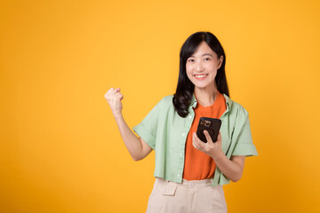 Excited young Asian woman in her 30s, wearing orange shirt and green jumper, using smartphone with fist up gesture on yellow studio background. Great news from mobile concept.