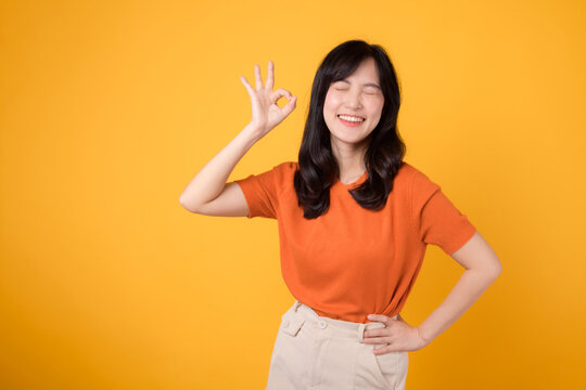 Expressing positivity, Asian woman 30s wears orange shirt, exhibits okay sign on vibrant yellow background. Hands gesture concept.