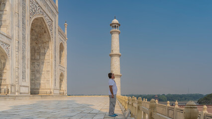Fototapeta na wymiar A man stands next to the Taj Mahal at the fence, looking into the distance. The hand is raised to the eyes. A white marble mausoleum and a high minaret against the blue sky. India. Agra.