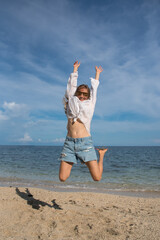 A happy blond woman with sunglasses is jumping on the beach. Attractive girl with long hair is wearing a denim shorts and a white shirt. Holidays and travel to other countries