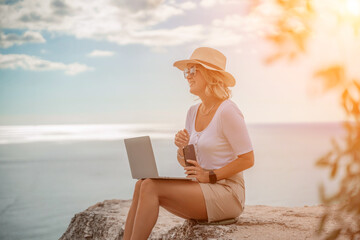 Freelance women sea working on the computer. Good looking middle aged woman typing on a laptop keyboard outdoors with a beautiful sea view. The concept of remote work.