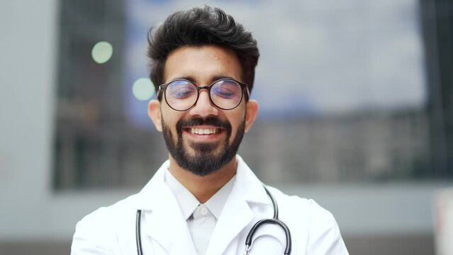 Close up portrait of a young smiling doctor in a white coat and glasses standing outside in front of a hospital building. Positive medical worker physician with stethoscope posing looking at camera