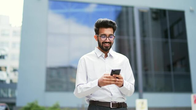 A young smiling businessman is using a smartphone while walking along the street near an office building. Happy handsome bearded male texting, checking email, chatting online, browsing social med