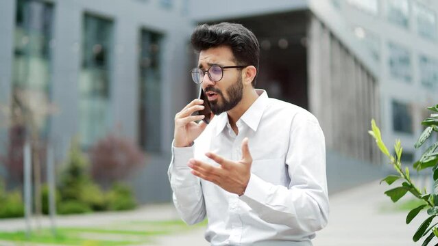 Angry young entrepreneur arguing while talking on smartphone sitting on a bench on the street near an office building. Excited, dissatisfied boss shouts into the phone, quarrels, expresses a problem