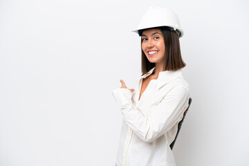 Young architect woman with helmet and holding blueprints isolated on white background pointing back