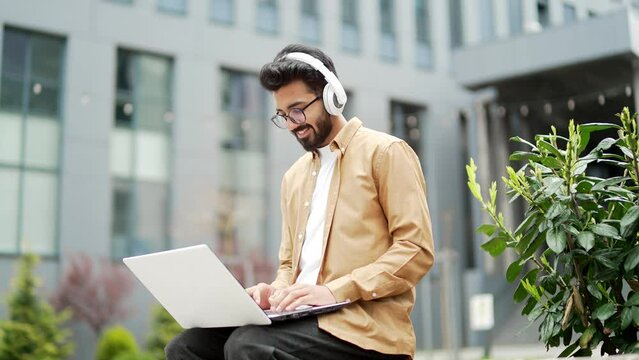 Young employee in headphones works on a laptop while sitting on a bench near the office building. Smiling handsome freelancer is chatting online with a client. IT specialist programmer uses a computer
