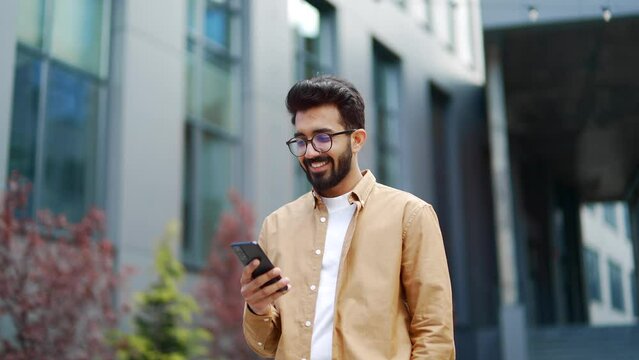A young smiling businessman is using a smartphone while walking along the street near an office building. Happy handsome bearded male checking email, chatting online, browsing social media, texting