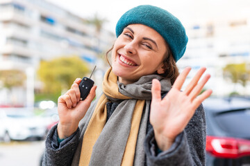 Brunette woman holding car keys at outdoors saluting with hand with happy expression