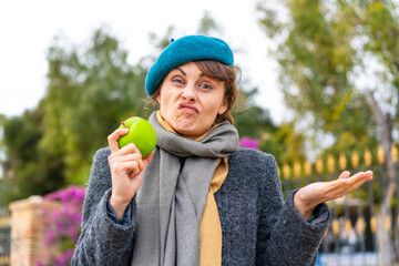 Brunette woman with an apple at outdoors making doubts gesture while lifting the shoulders