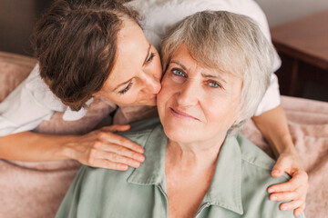 Fototapeta premium Adult girl child kiss overjoyed middle-aged mom relaxing together on sofa in living room