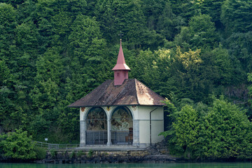 Famous Tell's Chapel at lakeshore of Lake Uri on a sunny spring day with woodland in the background. Photo taken May 22nd, 2023, Sisikon, Canton Uri, Switzerland. - obrazy, fototapety, plakaty