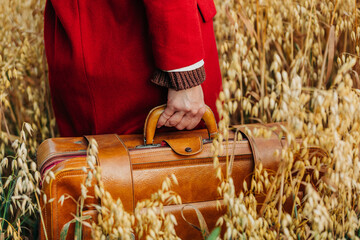 Fototapeta na wymiar Closeup view on woman in red coat hold vintage suitcase in oat field