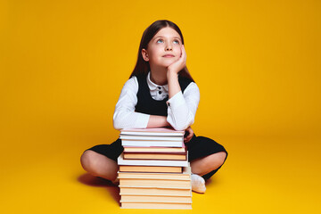 Thoughtful teenage girl sitting on floor with pile of books, holding hand under the chin and...