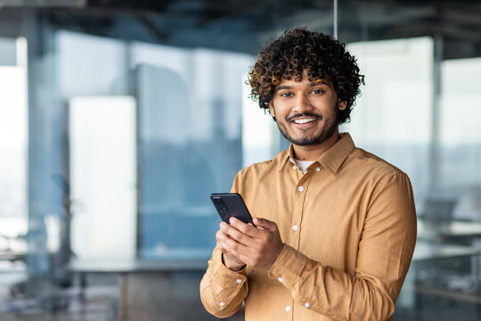 Portrait Of A Young Businessman Inside The Office, Hispanic Man Is Smiling And Looking At The Camera, The Man Is Holding A Phone In His Hands, Typing A Message And Browsing The Internet.