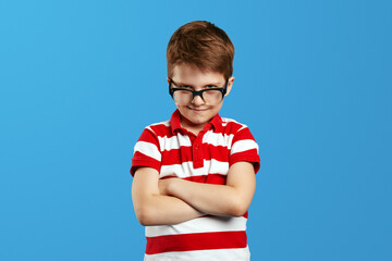 Serious kid in eyeglasses looking at camera. Portrait of focused little boy wearing spectacles and looking at camera with crossed hands, isolated on blue background.