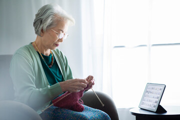 窓際のソファーに座って編み物をする高齢女性