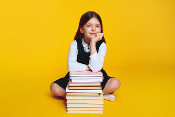 Elementary girl in school uniform, sitting near pile of books with crossed legs and hand under chin, isolated over yellow background