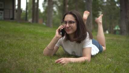 A young woman lies on the grass in the park and chats on the phone. Girl on the grass with a phone in glasses.