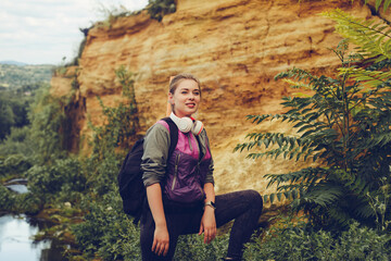 Naklejka na ściany i meble A girl on a hike near a river and a limestone wall with a backpack and headphones looks at the camera.