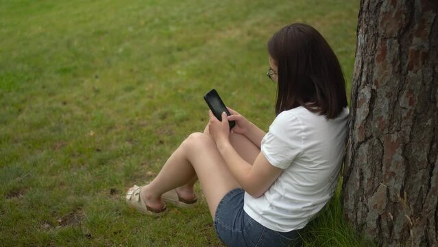 A young woman sits on the grass under a tree and chats on her smartphone. A girl with glasses is resting in the park with a phone.