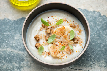 Bowl of cauliflower cream-soup topped with fried florets and croutons, horizontal shot on a grey and beige granite background