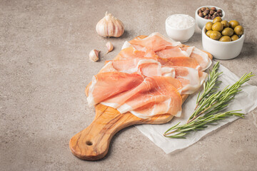 Thin slices of Italian prosciutto with green olives, spices and rosemary on wooden cutting broad. Raw dried meat.