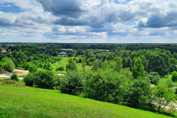 Fototapeta na wymiar Country landscape. Rural tourism. View from the hill to houses in forest against background of cloudy sky on summer day. European countryside, Poland. Concept of environmentally friendly city.
