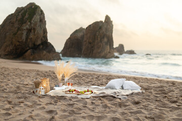 Fototapeta na wymiar Picnic at sunset with wine, glasses, snacks and fruits on blanket, candles and decor on sand, beautiful view on ocean