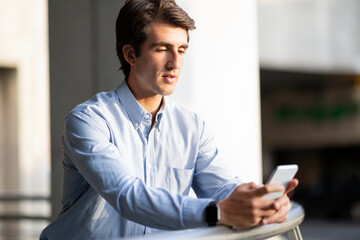 Millennial guy entrepreneur standing at office building, using smartphone