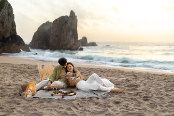 Romantic spouses having picnic, sitting on blanket and embracing, enjoying date on beach, celebrating romantic moment