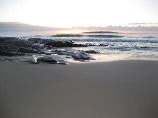 Just before dawn over the horizon with waves and rocks in the foreground and pristine sand.