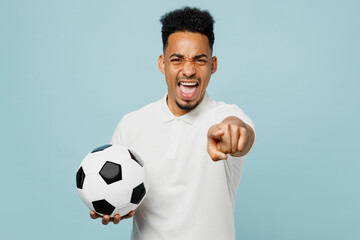 Young sad man fan wear basic t-shirt point index finger camera on you cheer up support football sport team hold in hand soccer ball watch tv live stream isolated on plain pastel blue color background.