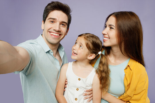Close up fun young parents mom dad with child kid daughter girl 6 year old wear blue yellow casual clothes do selfie shot pov mobile cell phone isolated on plain purple background Family day concept.