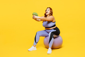 Full body sad hungry young chubby plus size big fat fit woman wear blue top warm up train sit on fit ball hold eat broccoli isolated on plain yellow background studio home gym. Workout sport concept.