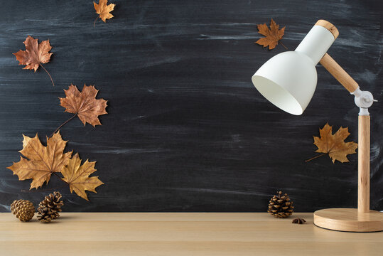Dive into the excitement of new school term with this captivating side-view picture of wooden desk with lamp and maple leaves on chalkboard background. Personalize copy-space with text or promotions
