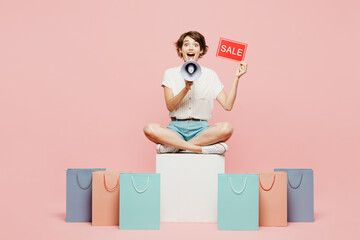 Full body young woman wear casual clothes hold shopping package bags,sale written text inscription sit scream in megaphone isolated on plain pink color background. Black Friday sale buy day concept.