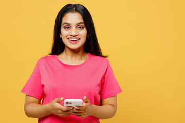 Young smiling cheerful happy Indian woman wear pink t-shirt casual clothes hold in hand use mobile cell phone browsing internet isolated on plain yellow background studio portrait. Lifestyle concept.