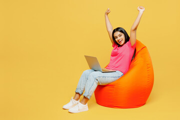Full body young happy IT Indian woman wearing pink t-shirt casual clothes sit in bag chair holding use work on laptop pc computer do winner gesture isolated on plain yellow background studio portrait.