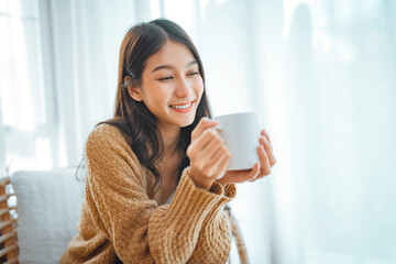 Happy asian woman relaxing drinking hot coffee or tea in holiday morning vacation on armchair at home, Cosy scene, Smiling pretty woman drinking hot tea in autumn winter.