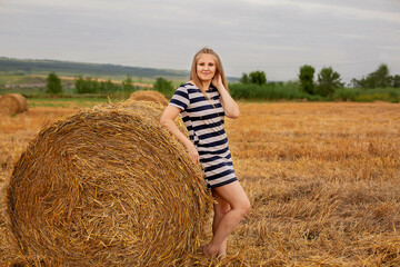 Naklejka na ściany i meble a long-haired blonde girl in a light summer dress is leaning against a haystack in a wheat field 